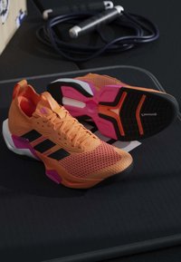 Chaussures de sport orange avec des rayures noires, des accents roses et des semelles texturées reposant sur un équipement d'entraînement sombre.