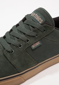 Tenisi din suede verde închis, cu șireturi plate, talpă din cauciuc și accente bej. Prezintă un mic etichetă cu logo pe lateral și detalii de cusătură.
