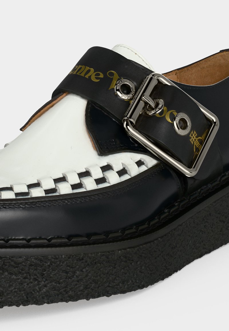 Schwarz-weißer Lederschuh mit dicker, strukturierter Sohle, großer silberner Schnalle, gewebtem weißen Detail und goldener Markenprägung auf schwarzem Riemen.