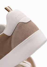 Beige mockaskicks med en vit läderhæl. Har en diskret logotyp, texturerade detaljer och en tjock vit sula.