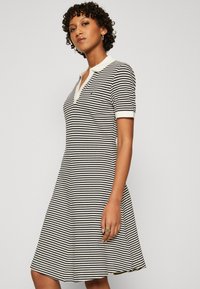 Gestreiftes Kleid in Schwarz und Weiß mit Polokragen, kurzen Ärmeln, ausgestelltem Schnitt, mit kontrastierenden strukturierten Akzenten und kleinem Logo.