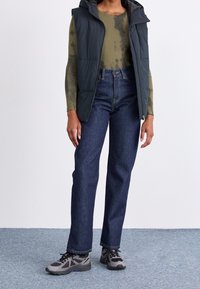 Dark green puffy vest over a tie-dye long-sleeve shirt, paired with blue straight-leg jeans and gray athletic shoes.