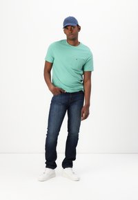 Mint green t-shirt with a pocket, dark blue jeans, and white sneakers. Male model stands with one hand on hip, wearing a blue cap.