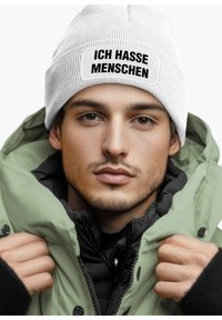 White ribbed beanie with a rectangular patch reading "ICH HASSE MENSCHEN" in bold black text, worn with a green puffer jacket.