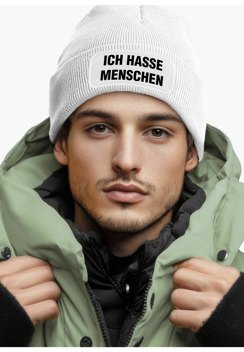 White ribbed beanie with a rectangular patch reading "ICH HASSE MENSCHEN" in bold black text, worn with a green puffer jacket.