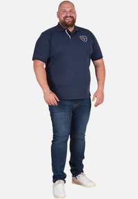 Polo shirt bleu marine à manches courtes, logo brodé sur la poitrine gauche, associé à un jean bleu foncé et des baskets blanches.
