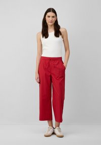 Pantaloni rossi svasati con vita elastica, tasche laterali e lunghezza cropped, abbinati a una canotta bianca e scarpe da ginnastica beige.