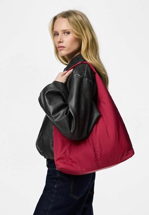 Femme blonde portant une veste noire et un jean foncé, portant un grand sac à bandoulière rouge, regardant par-dessus son épaule.