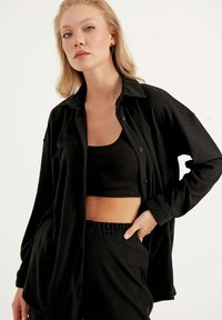 Schwarzes, geripptes Hemd mit Knöpfen und lockerer Passform, getragen über einem passenden schwarzen Tanktop und Bund. Weiche Textur, lässiges Design.