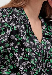 Schwarze Bluse mit floralem Muster, die kleine grüne und lila Blumen zeigt, mit weichem Material und V-Ausschnitt. Raffungen an den Schultern verleihen der Bluse Detail.