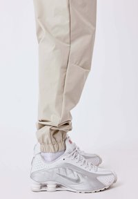 Persona que lleva pantalones jogger beige y zapatillas Nike blancas con suelas de aire visibles contra un fondo liso claro.