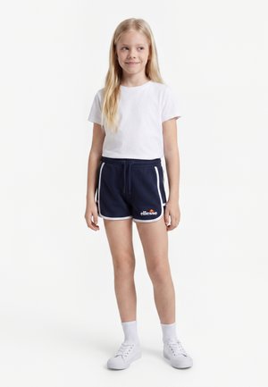 Młoda dziewczyna stoi, ubrana w białą koszulkę, granatowe krótkie spodenki sportowe z białym wykończeniem i logo, białe skarpetki i białe sneakersy na białym tle.