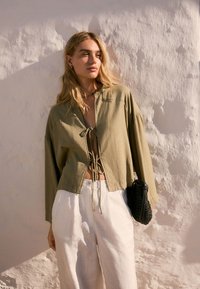 Khaki skrátená bunda so sťahovacou šnúrkou, dlhými rukávmi a voľným strihom, doplnená o biele široké nohavice a čiernu pletenú tašku.