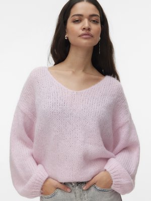 Γυναίκα με μακριά σκούρα μαλλιά που φοράει χαλαρό, ανοιχτό ροζ πλεκτό πουλόβερ με γυρισμένα μανίκια και ανοιχτόχρωμο τζιν, με τα χέρια στις τσέπες.