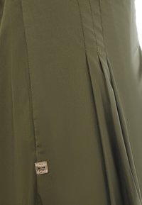 Gonna verde oliva scuro con pieghe e un’etichetta metallica in rilievo con la scritta "Manila Grace." Il tessuto sembra liscio con una leggera lucentezza.