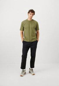 T-shirt vert olive à manches courtes, associée à un pantalon noir. Le modèle porte des baskets beige et gris avec des lacets et des semelles en caoutchouc.