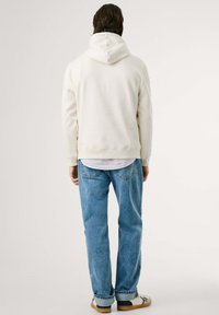 Licht beige capuchontrui met een zachte textuur, gecombineerd met lichtblauwe spijkerbroek en beige sneakers. Zichtbare achterkant.