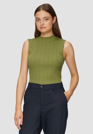 Femme aux longs cheveux bruns portant un haut en olive vert sans manches à côtes et un pantalon noir marine ajusté, se tenant avec une main dans sa poche.