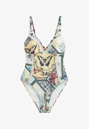 Femme portant un maillot de bain à motifs floraux et papillons et des lunettes de soleil, debout sur une rive rocheuse avec l'océan et le ciel en arrière-plan.