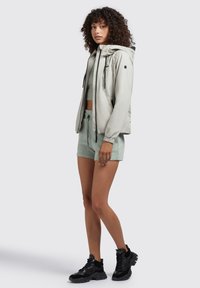  Lichtgrijze zip-up jas met capuchon, zwarte accenten en verstelbare koordjes, gecombineerd met een muntgroene crop top en bijpassende shorts. Zwarte sneakers.
