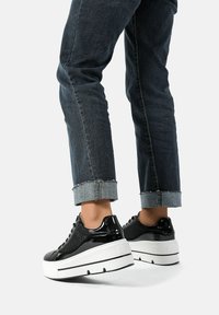 Sneakers neri in pelle lucida con una parte superiore testurizzata, caratterizzati da una suola bianca spessa e una striscia nera decorativa. Abbinati a jeans scuri di denim con risvolto.