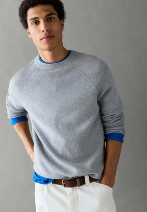 Jonge man met donker krullend haar, die een grijze trui draagt over een blauw overhemd en witte broek, staand met één hand in de zak tegen een grijze achtergrond.