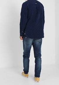 Tmavě modrá košile s bílými puntíky, zapnutým límečkem a zaobleným spodním lemem, spárovaná s modrými džínami a žlutými teniskami. Pohled zezadu.