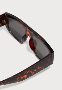 Schildpatt-Sonnenbrille mit dicken schwarz-orangenen gemusterten Rahmen, grauen Gläsern und rechteckiger Form. Mit integrierten Nasenpads.