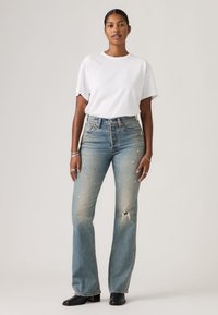 Γυναίκα όρθια, φορώντας λευκό μπλουζάκι, ψηλόμεσα φθαρμένα μπλε τζιν με τρύπα στο γόνατο, μαύρα μποτάκια στον αστράγαλο και ασημένια κοσμήματα σε απλό φόντο.