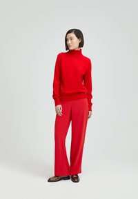 Roter Rollkragenpullover und weit geschnittene Hosen aus weichem Material. Auffällige Details sind die gerippten Bündchen und die glänzenden braunen Schuhe.