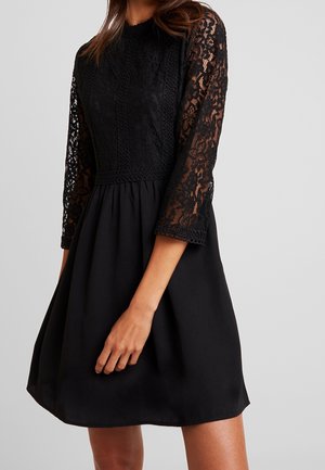 Vrouw draagt een zwarte jurk met kanten mouwen en lijfje, met een ronde halslijn en een uitlopende rok boven de knie.