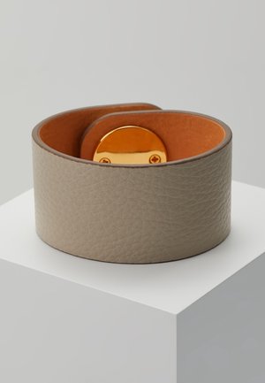 Large bracelet manchette en cuir taupe texturé avec intérieur lisse couleur fauve et fermoir ovale doré, posé sur un socle blanc.