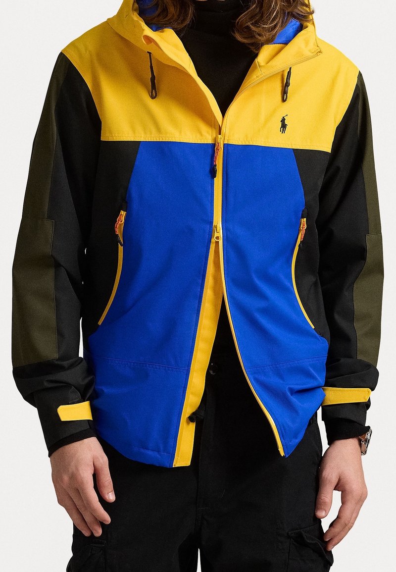 Persona che indossa una giacca color-block gialla, blu, nera e oliva con cerniera frontale e tasche laterali con cerniera, abbinata a pantaloni neri.