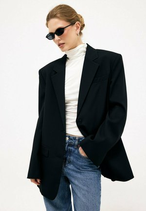 Ženska, oblečena v črno preveliko blazer, belo puli majico, modre kavbojke, z temnimi sončnimi očali in zvezanimi lasmi, stoji pred enobarvnim ozadjem.