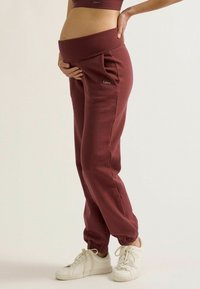 Bordeaux kleurige zwangerschap joggers met brede tailleband, frontzakken en elastische boorden. Gekocht met witte sneakers voor een casual look.