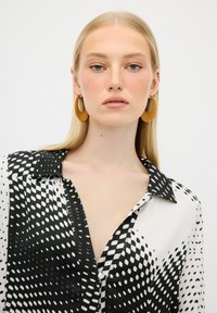 Blusa a collare bianca e nera con motivo a pois, caratterizzata da una morbida texture e una chiusura con bottoni; il modello indossa orecchini circolari gialli.
