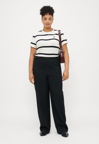 Valkoinen t-paita, jossa on mustia raitoja, lyhyet hihat ja pieni embleemi, yhdistettynä korkeavyötäröisiin mustiin housuihin ja mustiin liukuviin kenkiin.