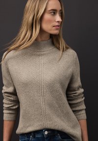 Pull beige côtelé avec un col montant, présentant des motifs diagonaux et des manches retroussées, associé à un jean bleu foncé.