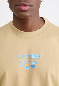 Šviesiai rudas trumpomis rankovėmis marškinėliai su mėlyna siuvinėta "AIR max" ir Nike logotipu, apvaliu kaklu ir lygia medvilnės tekstūra.