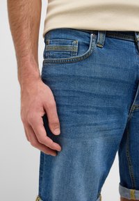Shorts de mezclilla en azul medio con un aspecto ligeramente desgastado, con costuras amarillas y puños doblados. Mano apoyada en el costado.