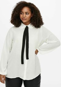 Blusa blanca con cuello festoneado y lazo negro. Mangas largas, corte holgado y dobladillo curvado. Tela suave y ligera.