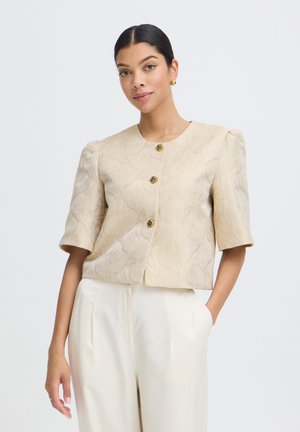 Femme portant une veste beige texturée à manches courtes avec des boutons dorés et un pantalon crème taille haute, debout devant un fond uni.