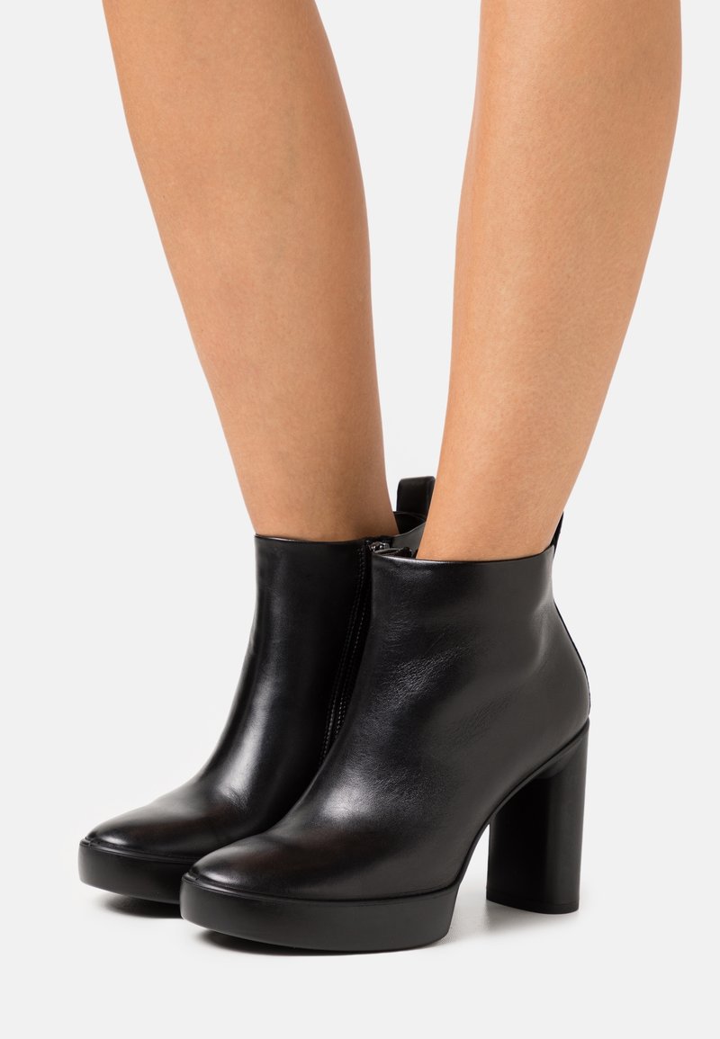 Bottines en cuir noir avec talons carrés hauts et fermetures éclair latérales portées sur jambes nues devant un fond clair uni.