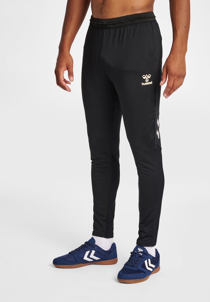 Man draagt zwarte hummel trainingsbroek en blauwe sneakers met witte chevron-logo's op een witte achtergrond.