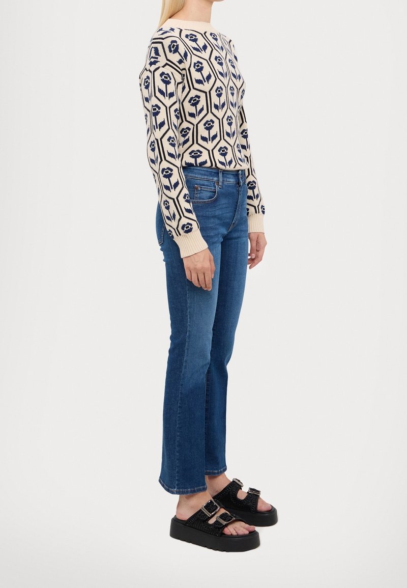 Femme portant un pull beige avec un motif floral bleu marine, un jean droit bleu et des sandales à plateforme noires avec des boucles, debout de profil.