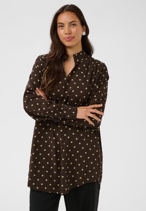 Femme aux longs cheveux bruns portant un chemisier marron à pois crème, les bras croisés, debout devant un fond uni clair.