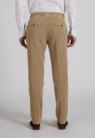 Achteraanzicht van een persoon die beige chino's draagt met knoopsluitingen op de achterzakken, een wit overhemd met lange mouwen en donkerbruine schoenen tegen een effen achtergrond.