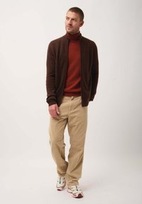 Cardigan in maglia marrone sopra un dolcevita rosso, abbinato a pantaloni in velluto a coste beige e sneakers bianche con dettagli rossi. Un look semplice e informale.