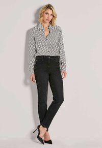 Vrouw met kort blond haar, gekleed in een gestreepte lange mouw shirt, zwarte high-waisted jeans en zwarte puntige hakken, staande tegen een eenvoudige muur.