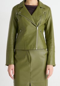 Femme portant une veste de motard en cuir vert olive avec des zips argentés et une jupe en cuir assortie, se tenant contre un fond blanc.